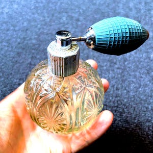 May include: A vintage perfume bottle with a clear, patterned glass body and a silver-colored metal cap. The atomizer bulb is teal and has a textured, grid-like pattern. The bottle is held in a hand.