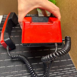 May include: A vintage red and black telephone with a coiled handset cord. The phone has a rectangular base and a separate handset. The handset and base are black, while the main body of the phone is red.