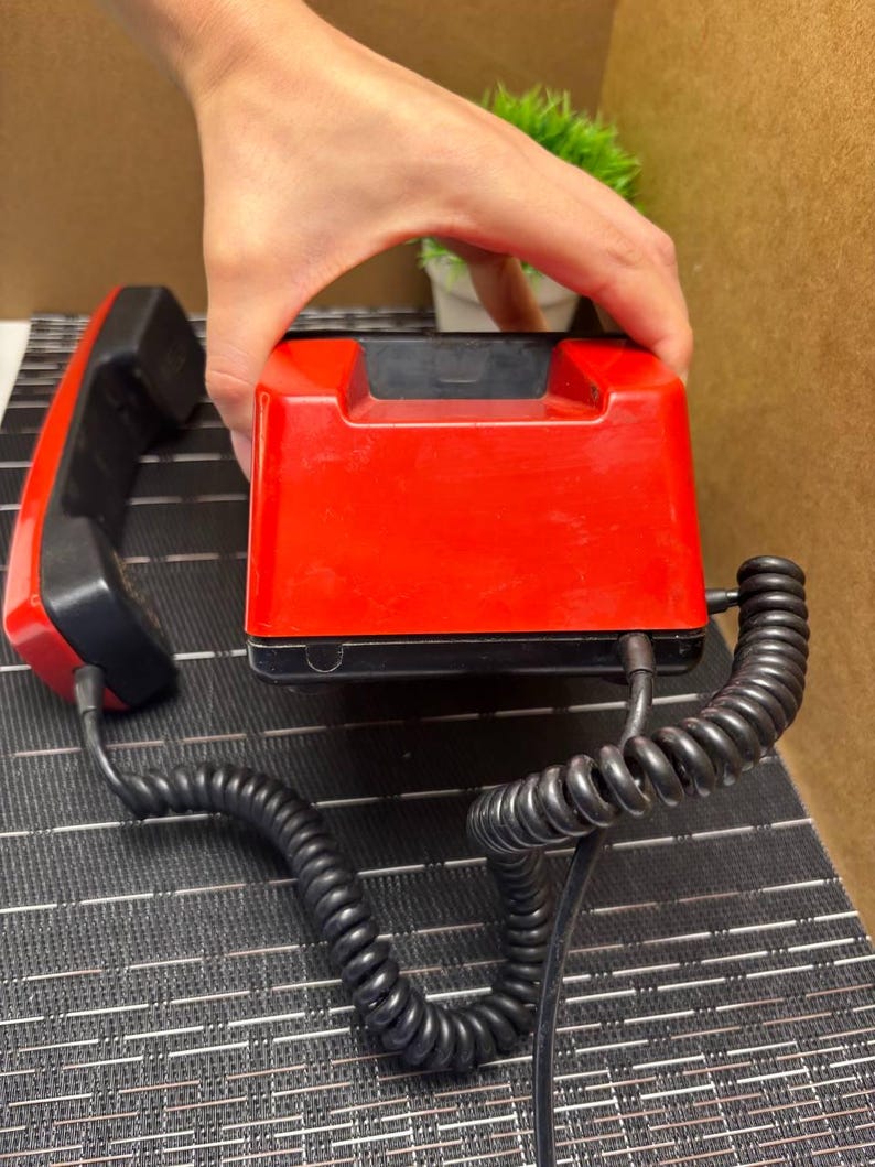May include: A vintage red and black telephone with a coiled handset cord. The phone has a rectangular base and a separate handset. The handset and base are black, while the main body of the phone is red.