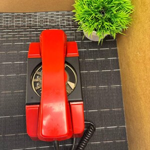 May include: A vintage red wall-mounted telephone with a black dial and base. The phone has a coiled handset cord and is set against a textured black surface. A small green plant in a white pot is in the background.