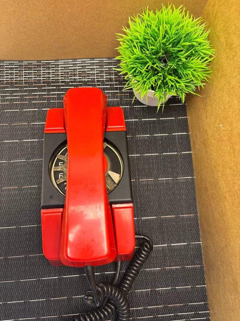 May include: A vintage red wall-mounted telephone with a black dial and base. The phone has a coiled handset cord and is set against a textured black surface. A small green plant in a white pot is in the background.