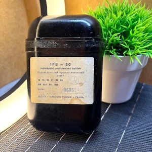 Op de afbeelding: Een zwarte, rechthoekige container met een label met tekst in een vreemde taal. Het label bevat de tekst "IPB-80" en andere details. Een kleine, groene plant in een witte pot is zichtbaar op de achtergrond.