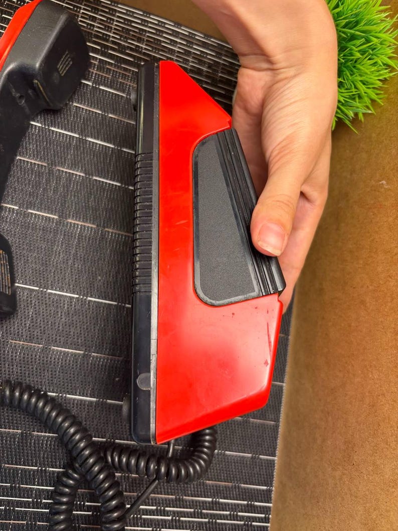 May include: A vintage red and black telephone receiver with a coiled cord. The receiver has a rectangular design with a black earpiece and a red body. The phone is likely from the 1980s or 1990s.