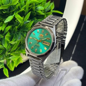 May include: A vintage Orient watch with a vibrant green dial and a silver-tone stainless steel band. The watch face displays the day and date, with gold-tone accents. The watch is displayed on a clear stand.