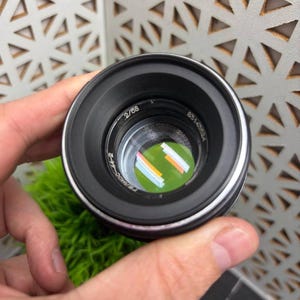 Peut inclure: Un objectif d'appareil photo noir avec des d&eacute;tails argent&eacute;s, portant les inscriptions "2/58" et "8314352A". L'objectif refl&egrave;te une image color&eacute;e. L'objectif est tenu dans une main, avec un fond vert textur&eacute;.