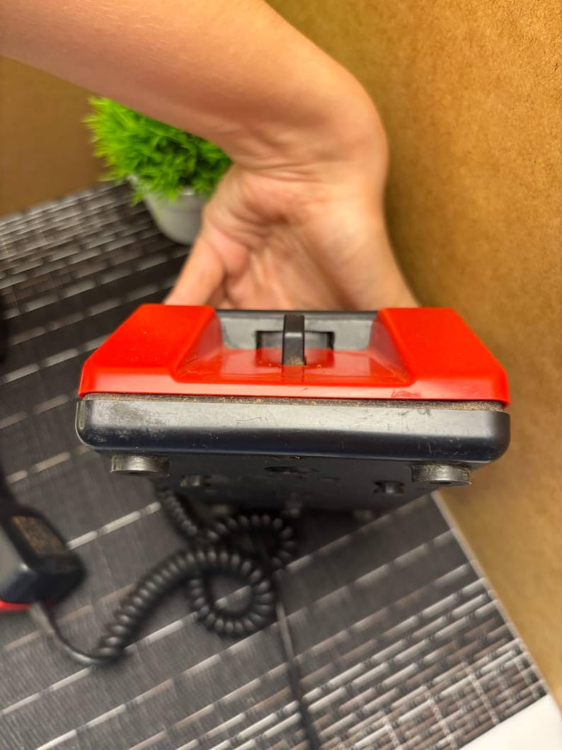 May include: A vintage black and red telephone with a coiled cord. The phone has a rectangular shape with a red top and a black base. The handset is not visible in the image.