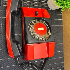 May include: A vintage red and black wall-mounted telephone with a rotary dial. The handset is black, and the phone's body is red with a black faceplate. The dial features numbers and a clear cover. A coiled black cord connects the handset to the phone.