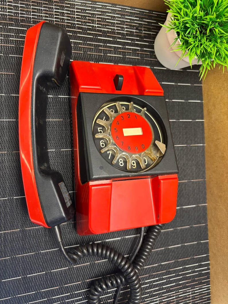 May include: A vintage red and black wall-mounted telephone with a rotary dial. The handset is black, and the phone's body is red with a black faceplate. The dial features numbers and a clear cover. A coiled black cord connects the handset to the phone.