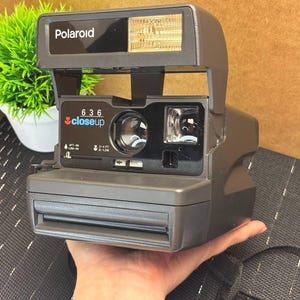 May include: A gray Polaroid 636 Closeup instant camera. The camera has a built-in flash and a lens with the word "closeup" printed on the front. The camera is held by a hand.
