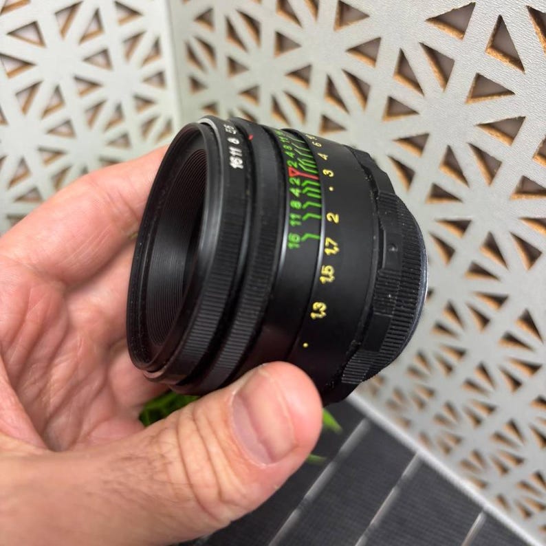 Peut inclure: Objectif d'appareil photo noir avec des indications de mise au point et d'ouverture. L'objectif comporte des marquages blancs, verts et rouges. L'objectif est tenu dans une main.