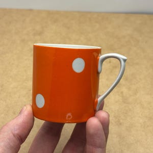 以下が含まれることがあります： 白い水玉模様と白い取っ手が付いた小さなオレンジ色のマグカップ