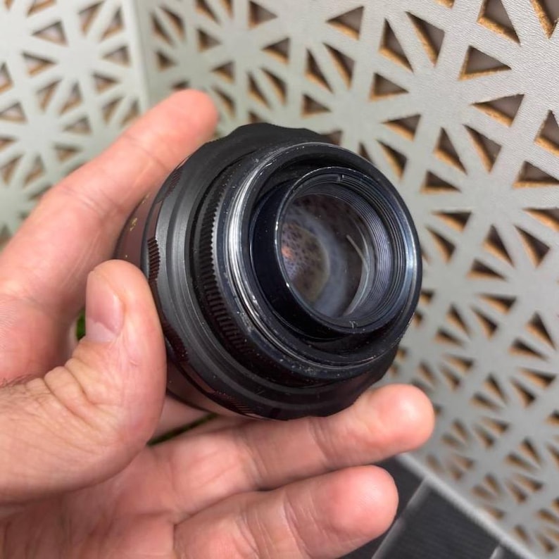 Peut inclure: Un objectif d'appareil photo noir avec une grande ouverture. L'objectif a une bague de mise au point rainur&eacute;e et un &eacute;l&eacute;ment en verre transparent. L'objectif est tenu dans une main.