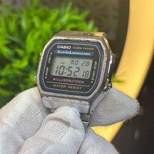May include: A silver-tone digital wristwatch with a rectangular face, featuring the text Casio, Alarm Chrono, Electro Luminescence, Illuminator, and Water Resist. The display shows the time 10:52 and the date MO 28. The metal casing shows visible wear.