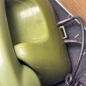 May include: An olive green rotary telephone featuring a curved handset resting in a cradle. A black coiled cord extends from the side of the base unit, which has a smooth, glossy plastic finish and visible surface scuffs on the handset.