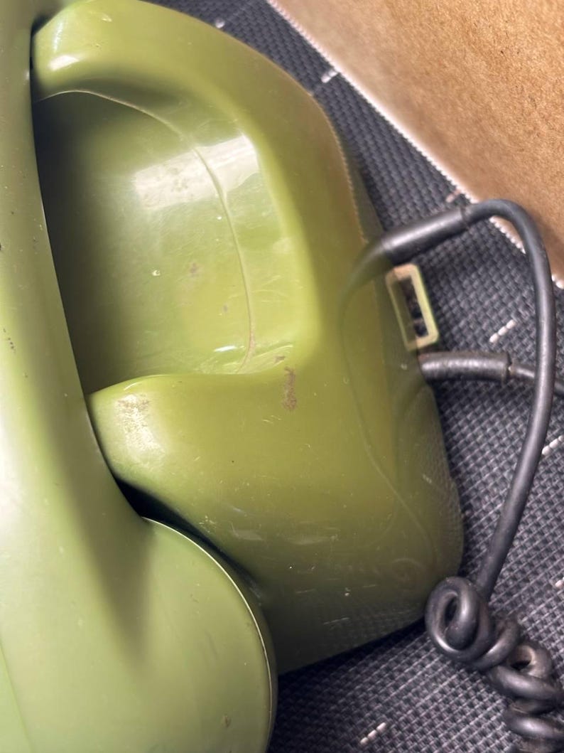 May include: An olive green rotary telephone featuring a curved handset resting in a cradle. A black coiled cord extends from the side of the base unit, which has a smooth, glossy plastic finish and visible surface scuffs on the handset.
