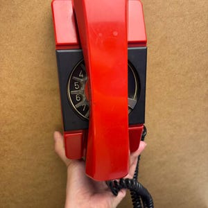 May include: A vintage red and black wall-mounted telephone. The handset is red, and the dial area is black with gold numbers. A black coiled cord extends from the base. The phone is held against a brown surface.