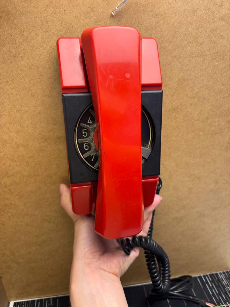 May include: A vintage red and black wall-mounted telephone. The handset is red, and the dial area is black with gold numbers. A black coiled cord extends from the base. The phone is held against a brown surface.