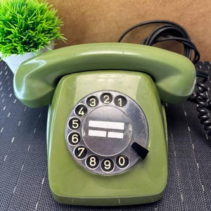 May include: An olive green rotary telephone featuring a circular dial with numbers zero through nine in black. The device includes a matching handset resting on top and a black coiled cord extending from the side, with a central white label area.