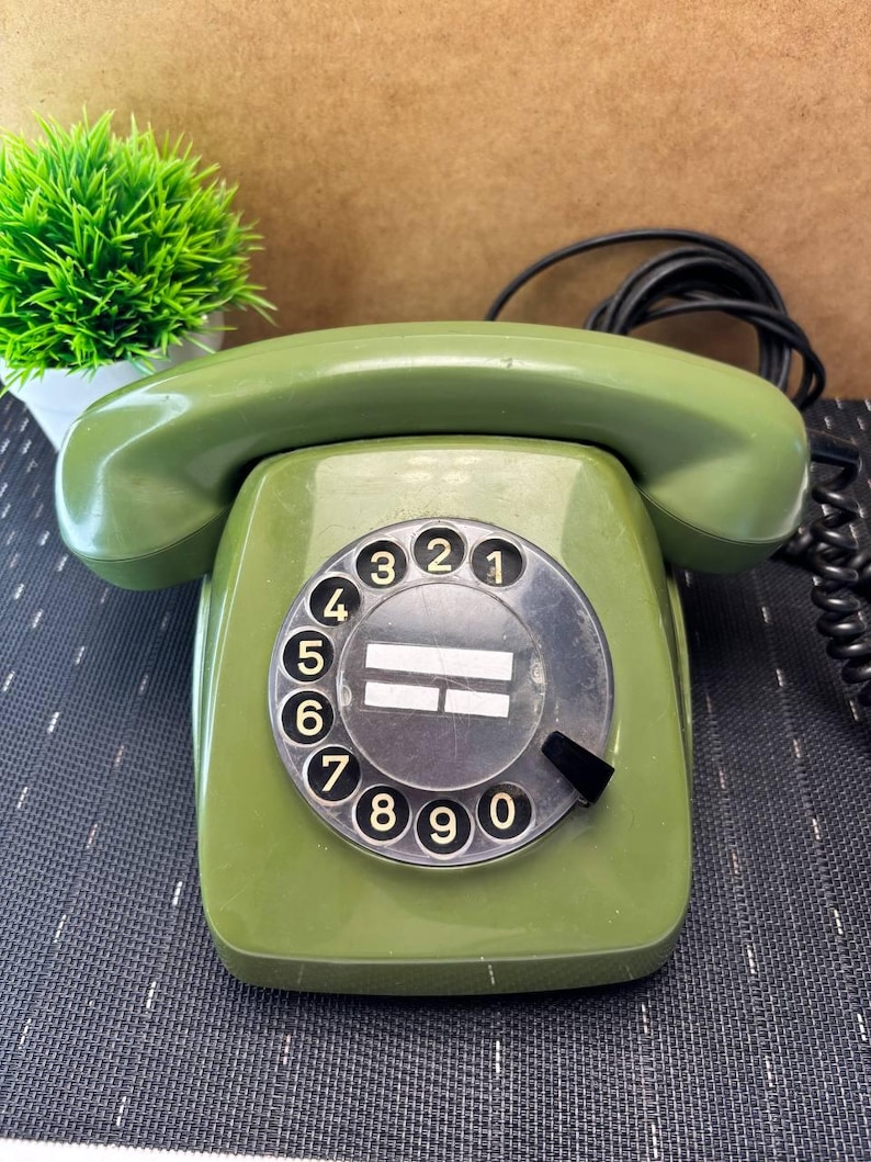 May include: An olive green rotary telephone featuring a circular dial with numbers zero through nine in black. The device includes a matching handset resting on top and a black coiled cord extending from the side, with a central white label area.