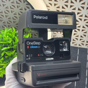 May include: A black Polaroid OneStep CloseUp instant camera. The camera has a built-in flash and a lens for close-up shots. The camera has the text "Polaroid" and "OneStep closeup" printed on it. The camera is designed to use 600 film.