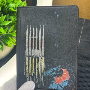 May include: Black notebook with a space-themed design. The cover features a row of rockets with yellow and white flames, a globe, and stars. The notebook is held by a gloved hand.