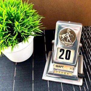 May include: A silver-toned desktop calendar with a horse and rider design. The calendar shows the date as March 20th and the day of the week as Wednesday.