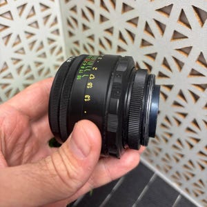 Peut inclure: Un objectif d'appareil photo noir avec des marquages verts, jaunes et blancs. L'objectif comporte une s&eacute;rie d'anneaux et de chiffres indiquant la distance focale et les r&eacute;glages d'ouverture. L'objectif est tenu dans une main.