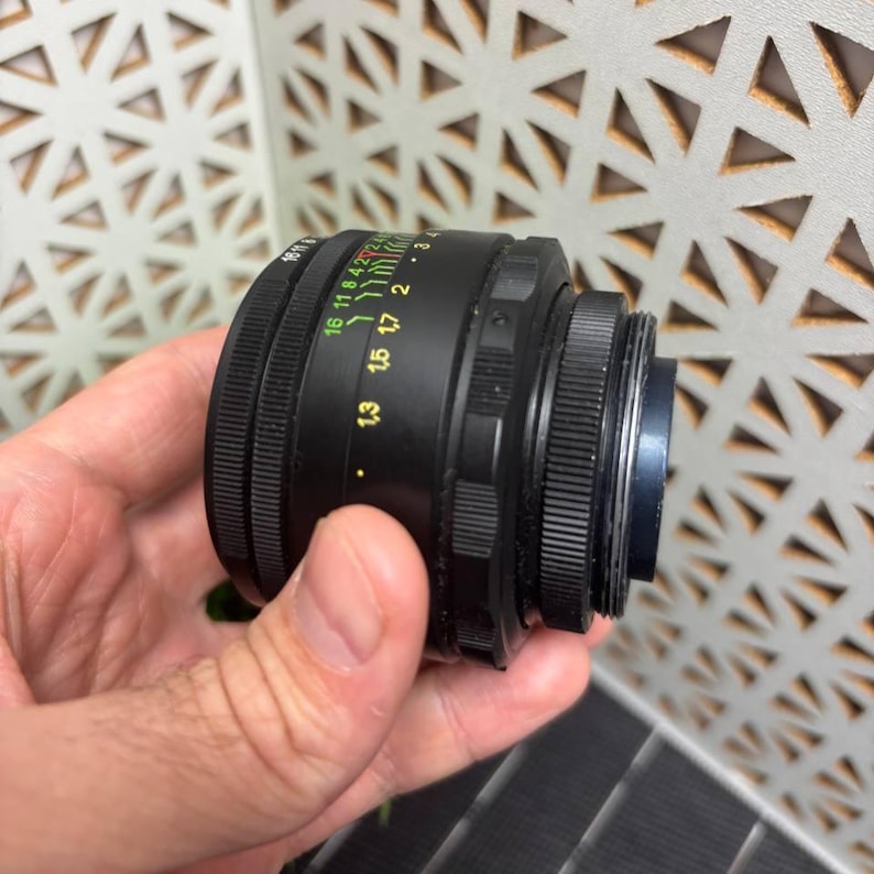 Peut inclure: Un objectif d'appareil photo noir avec des marquages verts, jaunes et blancs. L'objectif comporte une s&eacute;rie d'anneaux et de chiffres indiquant la distance focale et les r&eacute;glages d'ouverture. L'objectif est tenu dans une main.