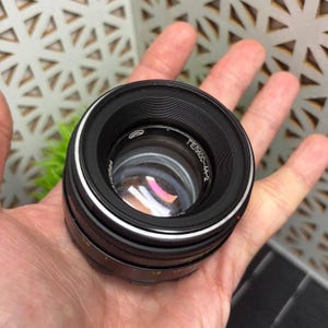 Peut inclure: Un objectif d'appareil photo noir avec un anneau argent&eacute; et le texte "Гелиос-44-2". L'objectif est tenu dans une main ouverte. L'objectif a une surface r&eacute;fl&eacute;chissante.