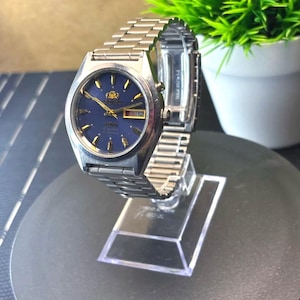 May include: A silver-toned wristwatch with a blue face and gold accents. The watch has a metal band and is displayed on a clear stand. The watch face displays the brand name "Orient" and the day of the month.