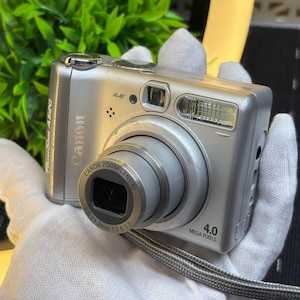 以下が含まれることがあります： 4.0メガピクセルのラベル、Canonズームレンズ4x、5.8-23.2mm 1:2.6-5.5のレンズ仕様を備えたシルバーのCanon PowerShot A520デジタルカメラ。このコンパクトカメラには、内蔵フラッシュ、AiAFセンサー、グレーのリストストラップが装備されています。