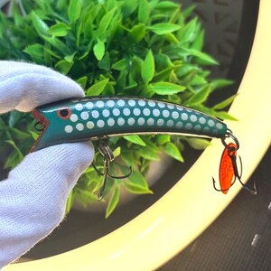 May include: A teal fishing lure with a white polka dot pattern, a painted red eye, and a metallic finish. The lure features two silver-toned treble hooks attached to the underside and tail, with a small orange translucent plastic blade attached.