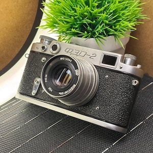 Puede incluir: Una cámara ФЭД-2 vintage con lente plateada y cuerpo negro texturizado. La cámara tiene el texto "ФЭД-2" y "N013594" impreso. Una pequeña planta verde en una maceta blanca se encuentra detrás de la cámara.