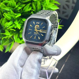 May include: A vintage Orient watch with a silver-tone stainless steel band and a square face. The watch face is dark blue with gold-tone hands and markers. The watch displays the day and date.