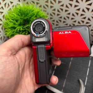 May include: A red and black Alba camcorder with 6x digital zoom. The camcorder is held in a hand and has the text "ALBA" and "FIELD CAMCORDER" printed on it. The lens has the text "f=2.8 f=8.5" printed on it.