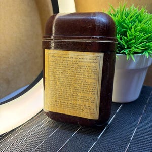 Op de afbeelding: Een donkerbruine, rechthoekige container met een afgeronde bovenkant en een label met tekst. De container staat op een donkere ondergrond, met een kleine groene plant in een witte pot op de achtergrond.