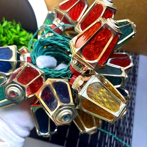 May include: A collection of small, multi-coloured lanterns with gold-coloured frames. The lanterns feature red, blue, green, and yellow stained glass panels. The string is green, and the lights are illuminated.