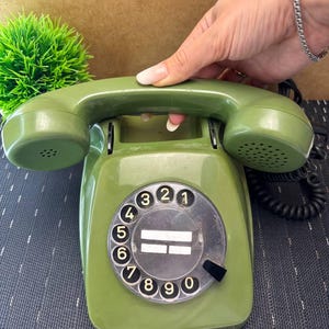 May include: An olive green rotary telephone featuring a circular dial with black numbers zero through nine and a matching handset resting on top. The device includes a black coiled cord and a central circular area with two white horizontal lines.