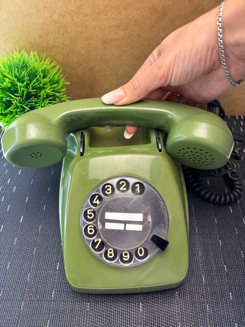 May include: An olive green rotary telephone featuring a circular dial with black numbers zero through nine and a matching handset resting on top. The device includes a black coiled cord and a central circular area with two white horizontal lines.