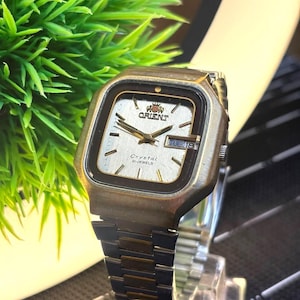 May include: A vintage Orient Crystal 21 Jewels watch with a silver dial and a rectangular gold-tone case. The watch features a day and date display and a two-tone metal band. The watch face displays the Orient logo.