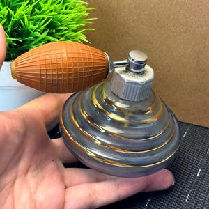 May include: Vintage perfume bottle with a silver and gold design. The bottle has a bulb-shaped atomizer with a brown, textured rubber bulb. The bottle's body is a rounded shape with gold bands.