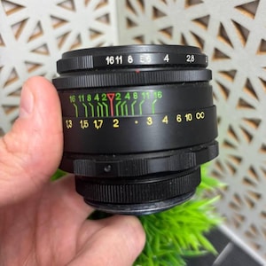 Peut inclure: Un objectif d'appareil photo noir avec des marquages blancs et jaunes. L'objectif comporte des chiffres et des mesures, notamment des ouvertures et des &eacute;chelles de distance. L'objectif est tenu dans une main.