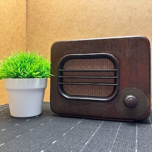 Puede incluir: Una radio de madera de estilo vintage, de color marrón oscuro, con una rejilla de altavoz y un botón de sintonización. A la izquierda de la radio hay una pequeña maceta blanca con hierba artificial verde brillante. La radio está sobre una superficie gris oscura.