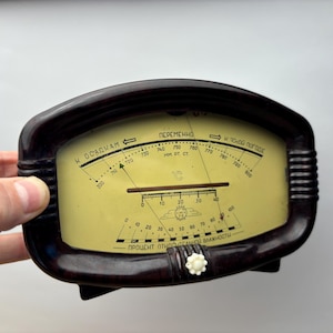 May include: A black and yellow analogue barometer with a needle pointing to the right. The barometer has a white knob on the bottom and the text "И ОСАДКАМ" and "ПЕРЕМЕННО" printed on the face.