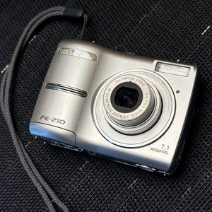 May include: A silver Olympus FE-210 digital camera with a lens and flash. The camera has a 7.1 megapixel resolution and a zoom lens. A grey strap is attached. The camera is on a black textured surface.