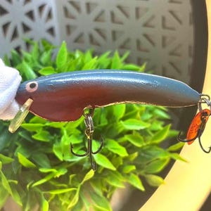 May include: A fishing lure with a smooth, curved body featuring a dark blue top and a reddish-brown underside. The lure includes a small white eye, a clear plastic diving lip at the front, and two silver metal treble hooks attached to the bottom.