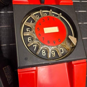 May include: A vintage rotary dial telephone with a red and black color scheme. The dial is red with white numbers and a clear finger wheel. The phone has a black body and a red receiver cradle. The phone is a classic design.