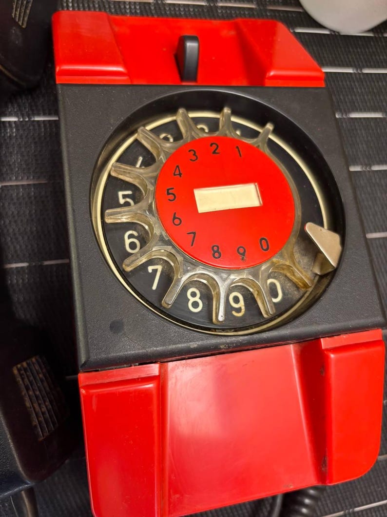 May include: A vintage rotary dial telephone with a red and black color scheme. The dial is red with white numbers and a clear finger wheel. The phone has a black body and a red receiver cradle. The phone is a classic design.