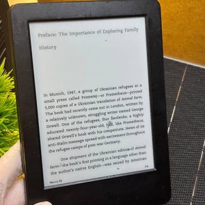 Könnte beinhalten: Ein schwarzer E-Reader, der Text aus einem Buch mit dem Titel "Vorwort: Die Bedeutung der Erforschung der Familiengeschichte" anzeigt. Der Bildschirm zeigt Text über eine ukrainische Übersetzung von Animal Farm. Der E-Reader wird in einer Hand gehalten.