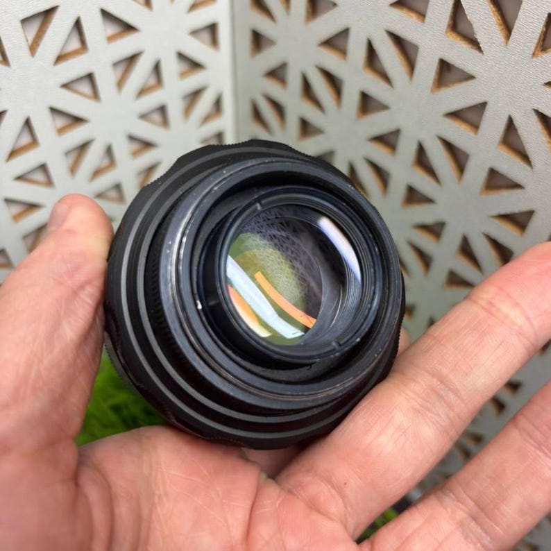 Peut inclure: Un objectif d'appareil photo noir avec un &eacute;l&eacute;ment en verre transparent, tenu dans une main. L'objectif a une bague de mise au point stri&eacute;e et une surface r&eacute;fl&eacute;chissante, capturant la lumi&egrave;re et les couleurs. L'arri&egrave;re-plan est une surface claire avec un motif g&eacute;om&eacute;trique.